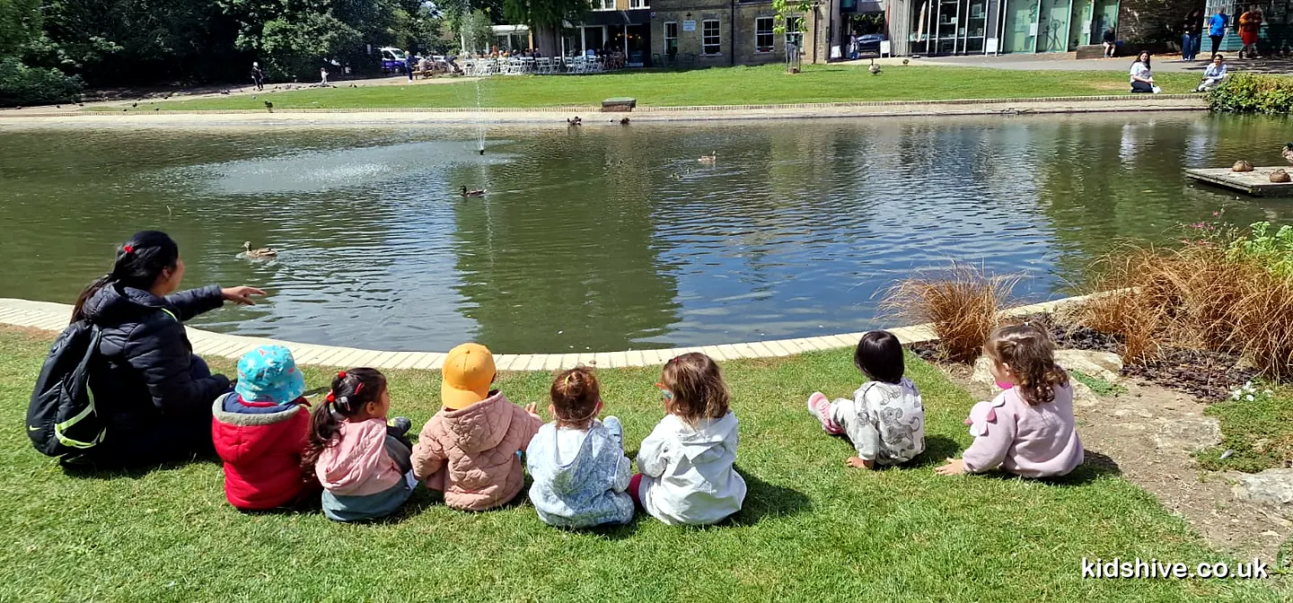 A childminder and seven toddlers sitting on the grass beside a park pond, watching the ducks
